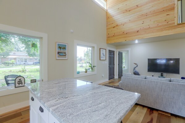 A modern living room with a large kitchen island, a grey sofa, and a flat-screen TV. - Blue Heron - New Modern Home in Carrabelle (Carrabelle)