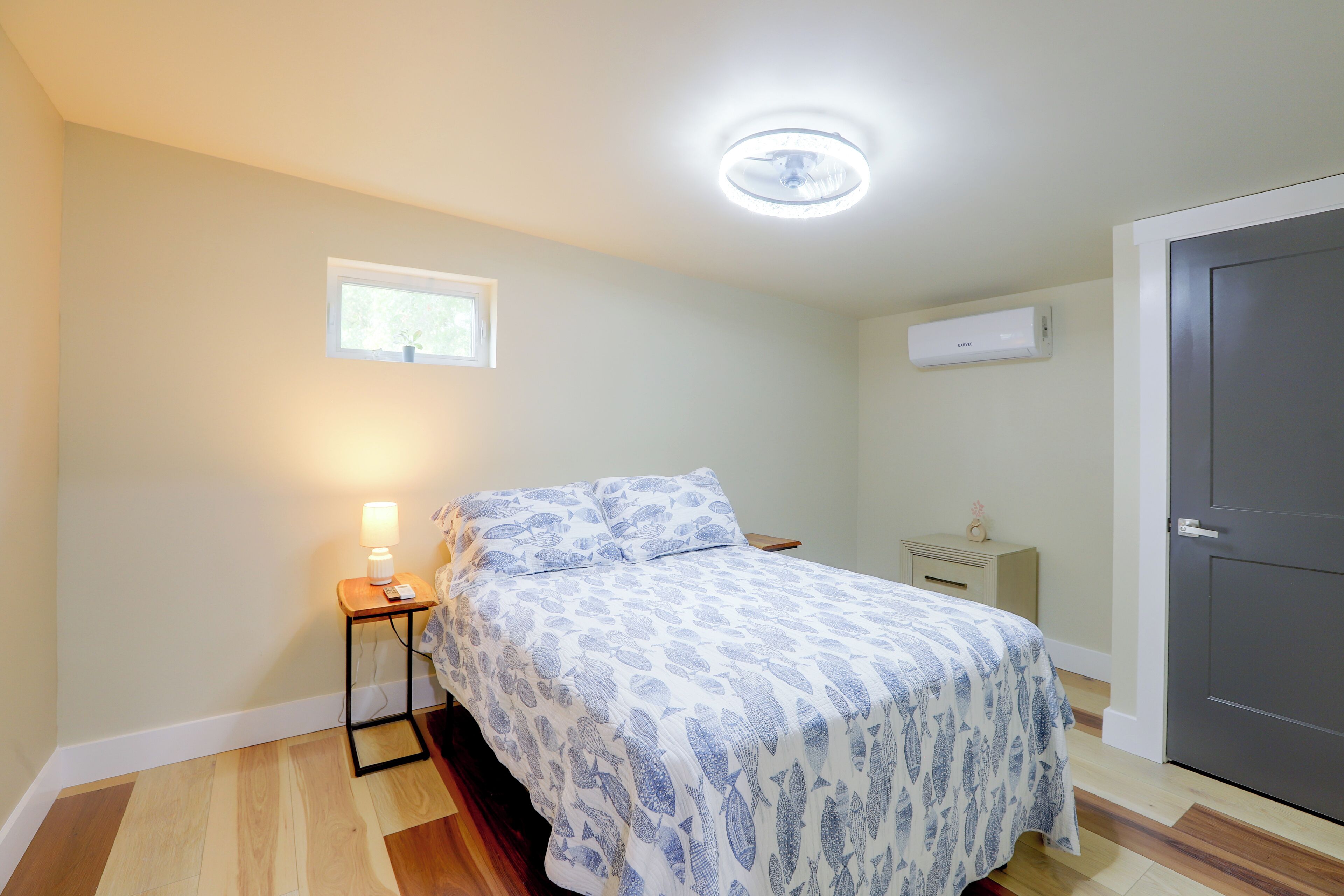 Ein Schlafzimmer mit Bett, Nachttisch, Lampe, Fenster, Klimaanlage und einer Tür.