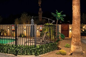 Área externa bem iluminada, com piscina, cerca de metal e diversas plantas.