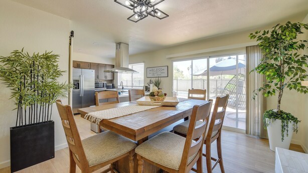 A dining area with a wooden table and chairs, a modern kitchen in the background, and a large window with a view of a house. - Private Pool Retreat Near Tucson Attractions (Tucson)