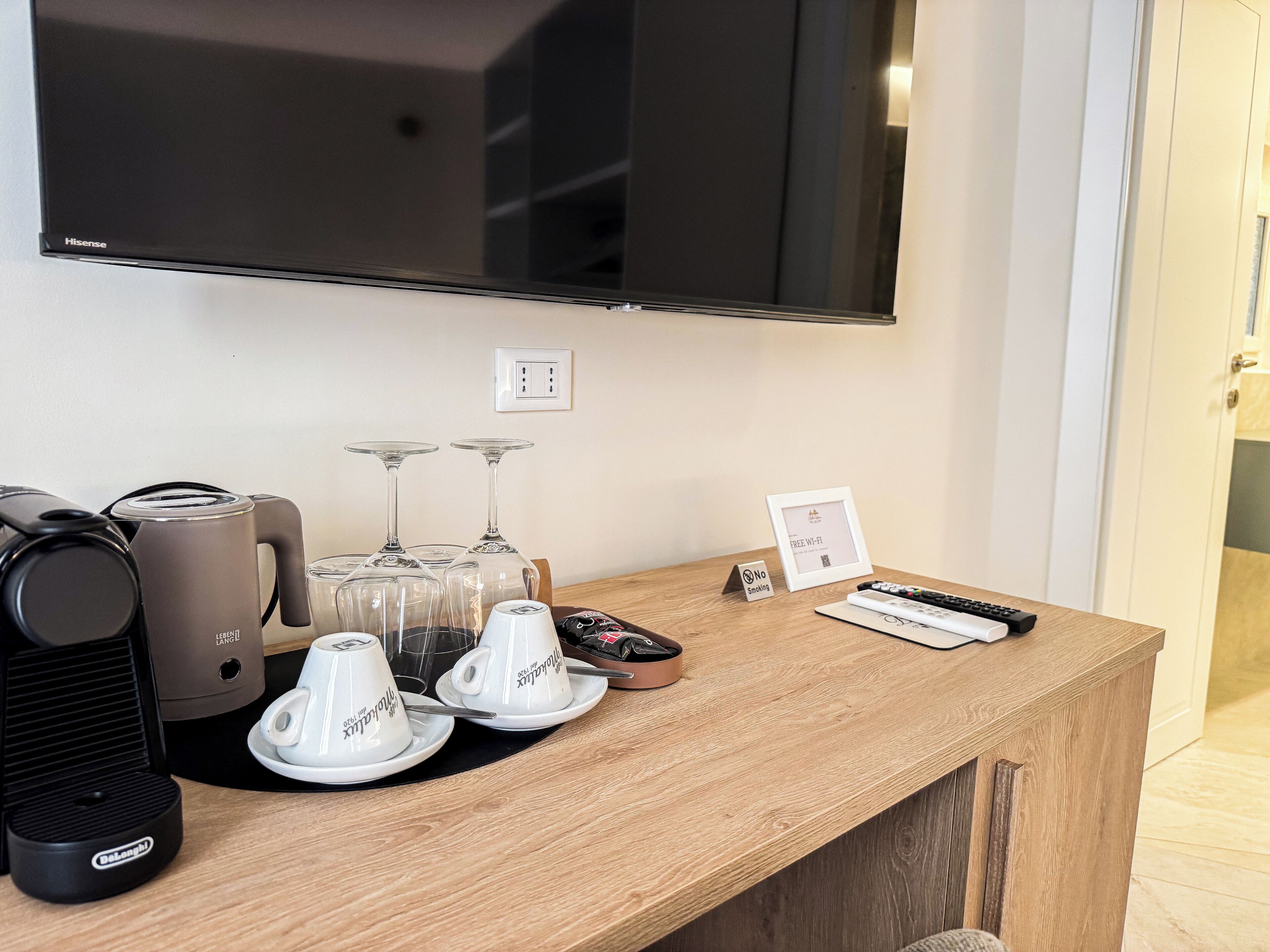Habitación de hotel con televisión de pantalla plana montada en la pared, una mesa de madera con una cafetera, dos copas de vino y un control remoto.