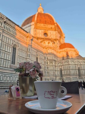 A cup with 'I'Opera Caffè' text on a saucer, a vase with flowers, and a view of a large building with a dome. - Bella Vigna (Florence)
