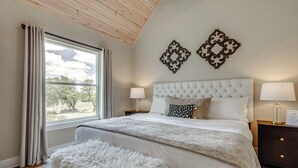 A bedroom with a large window, a tufted headboard, two decorative wall pieces, a bedside lamp, and a fluffy white bench. - Hot Tub & Fire Pit! Texas Wine Country Retreat (Fredericksburg)