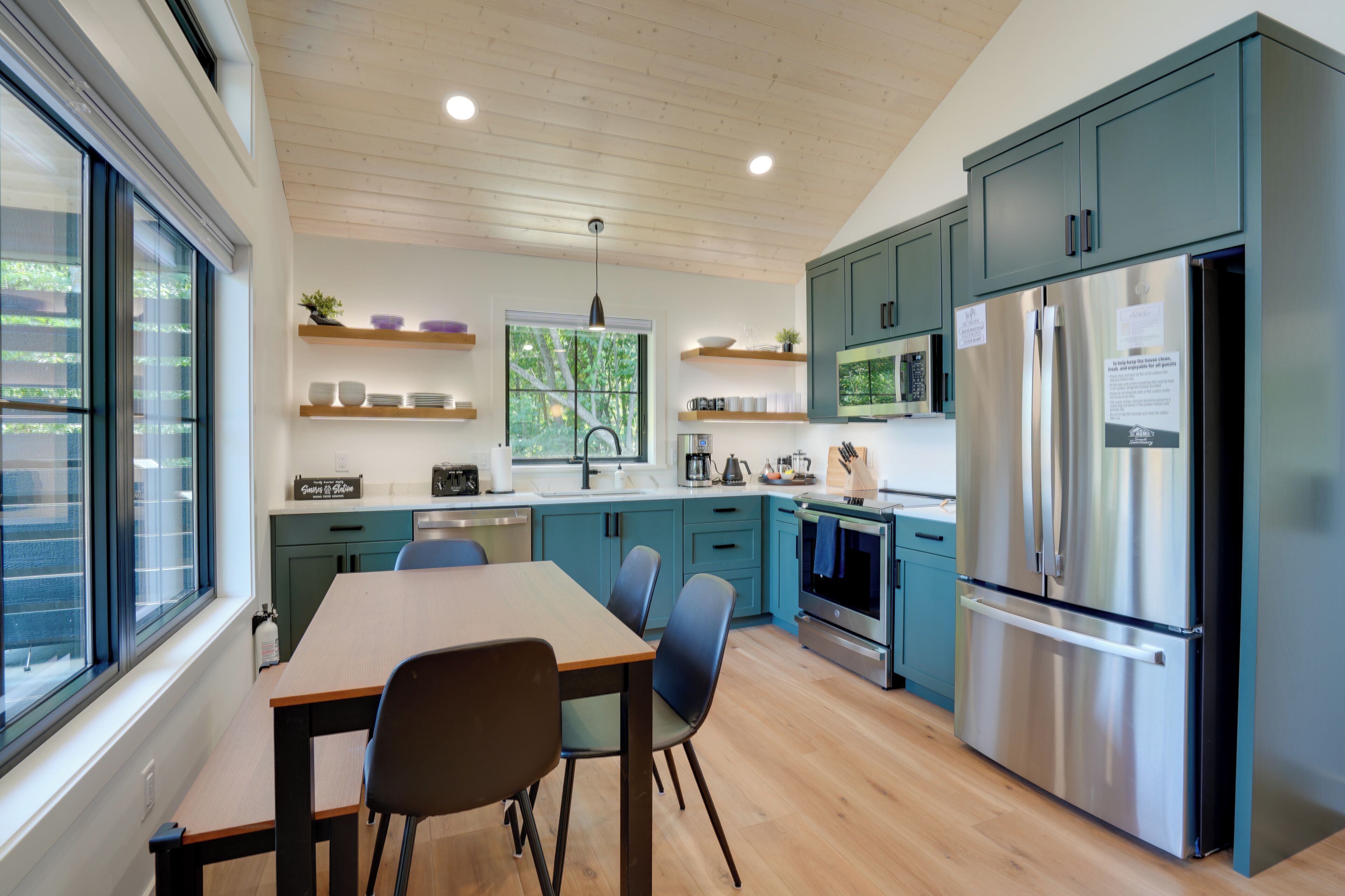 A modern kitchen with teal cabinets, stainless steel appliances, and a dining area with a wooden table and chairs.