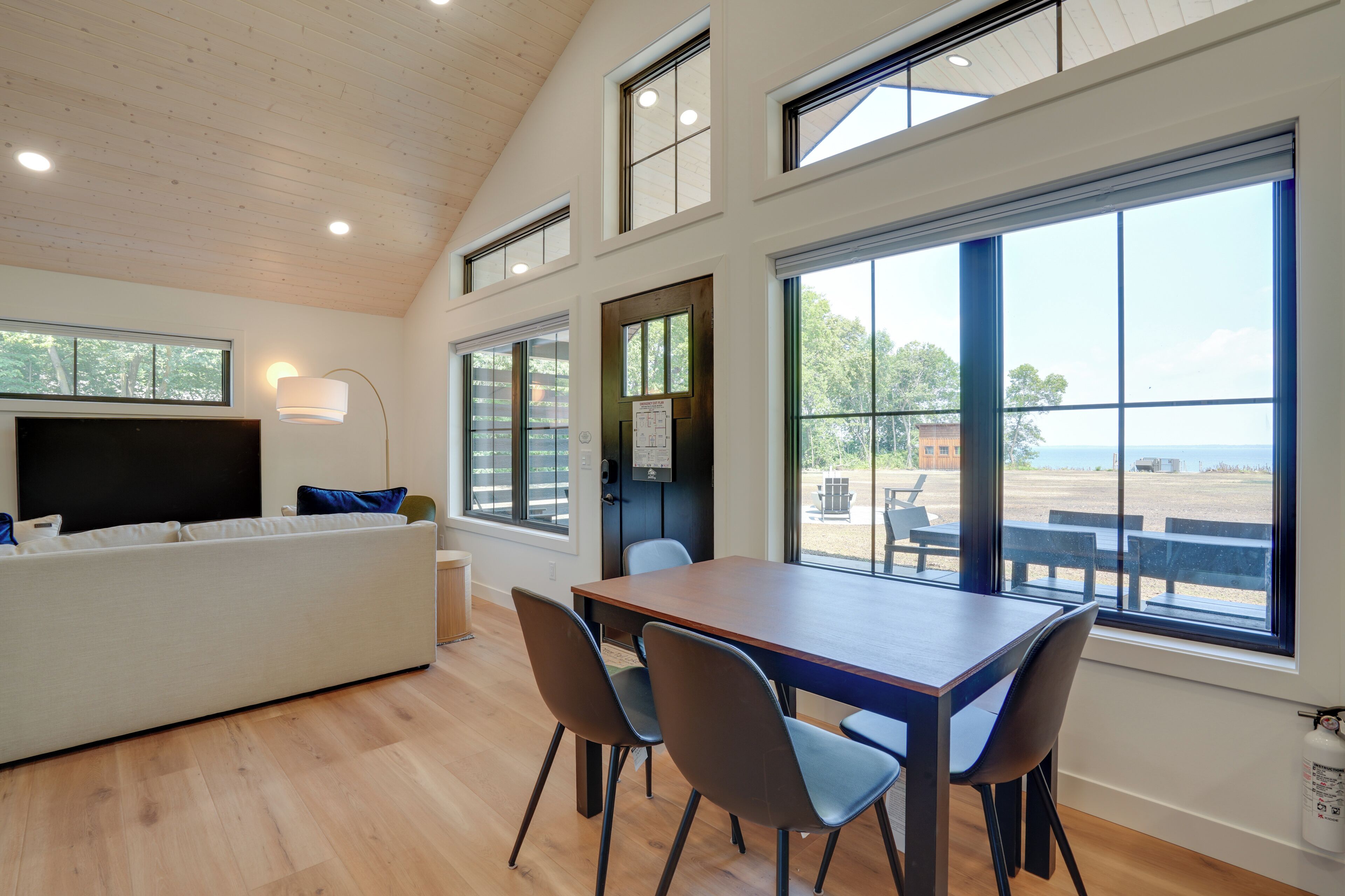 A modern interior with a wooden floor, a white sofa, a dining table with chairs, and large windows offering a view of the outdoors.