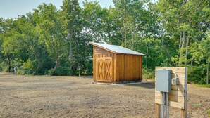 A wooden shed with a metal roof in a grassy field, surrounded by trees.
