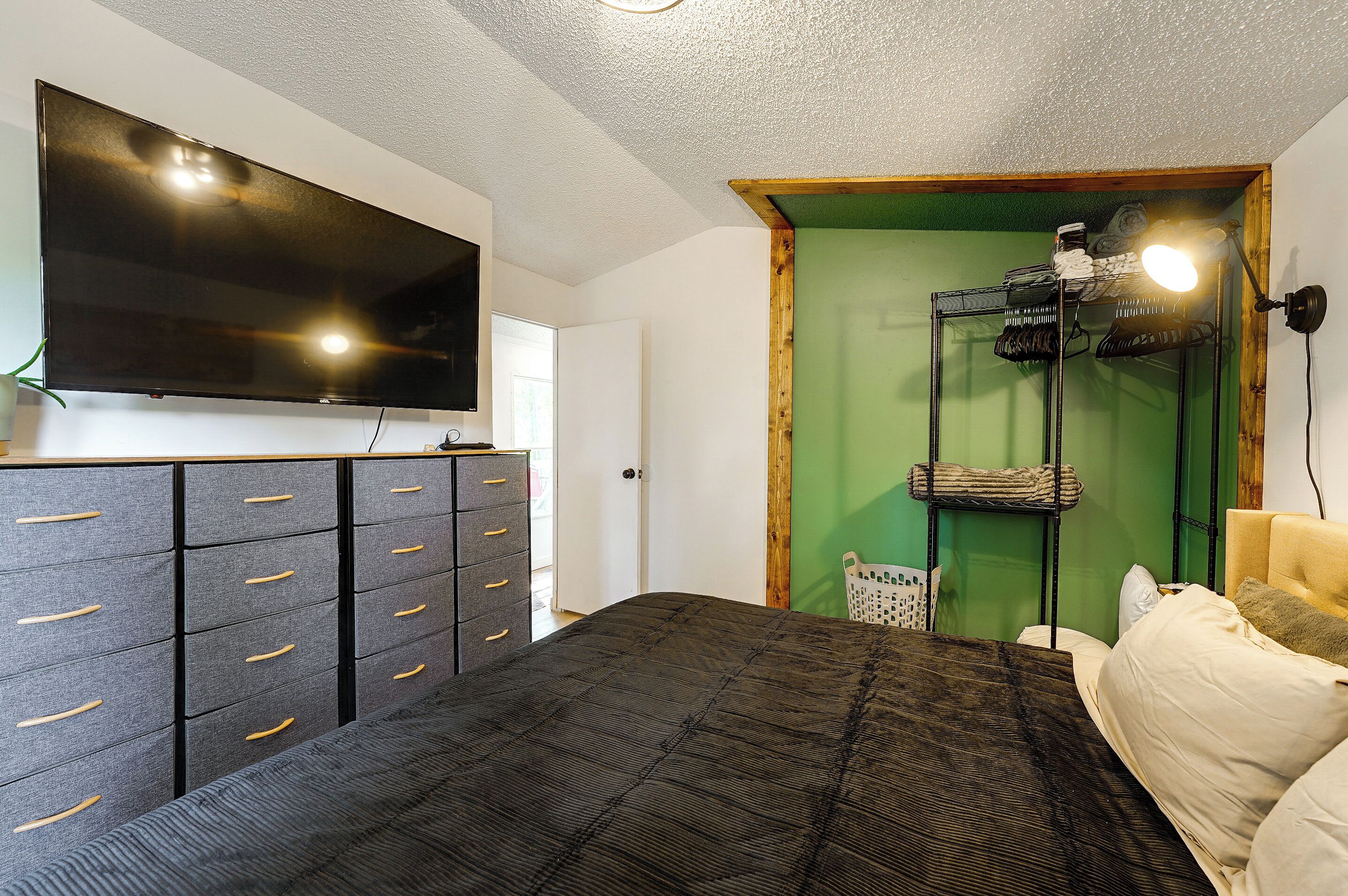 Une chambre à coucher avec un lit, une commode à tiroirs, un téléviseur fixé au mur et une porte verte.
