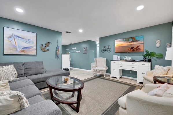 Uma sala de estar moderna com um sofá cinzento, uma mesa de centro redonda e uma poltrona branca. Há duas pinturas nas paredes: uma com um barco e outra com uma paisagem praiana.