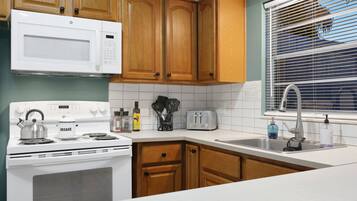 A kitchen with wooden cabinets, a white stove, a microwave, a toaster, and a window with blinds.
