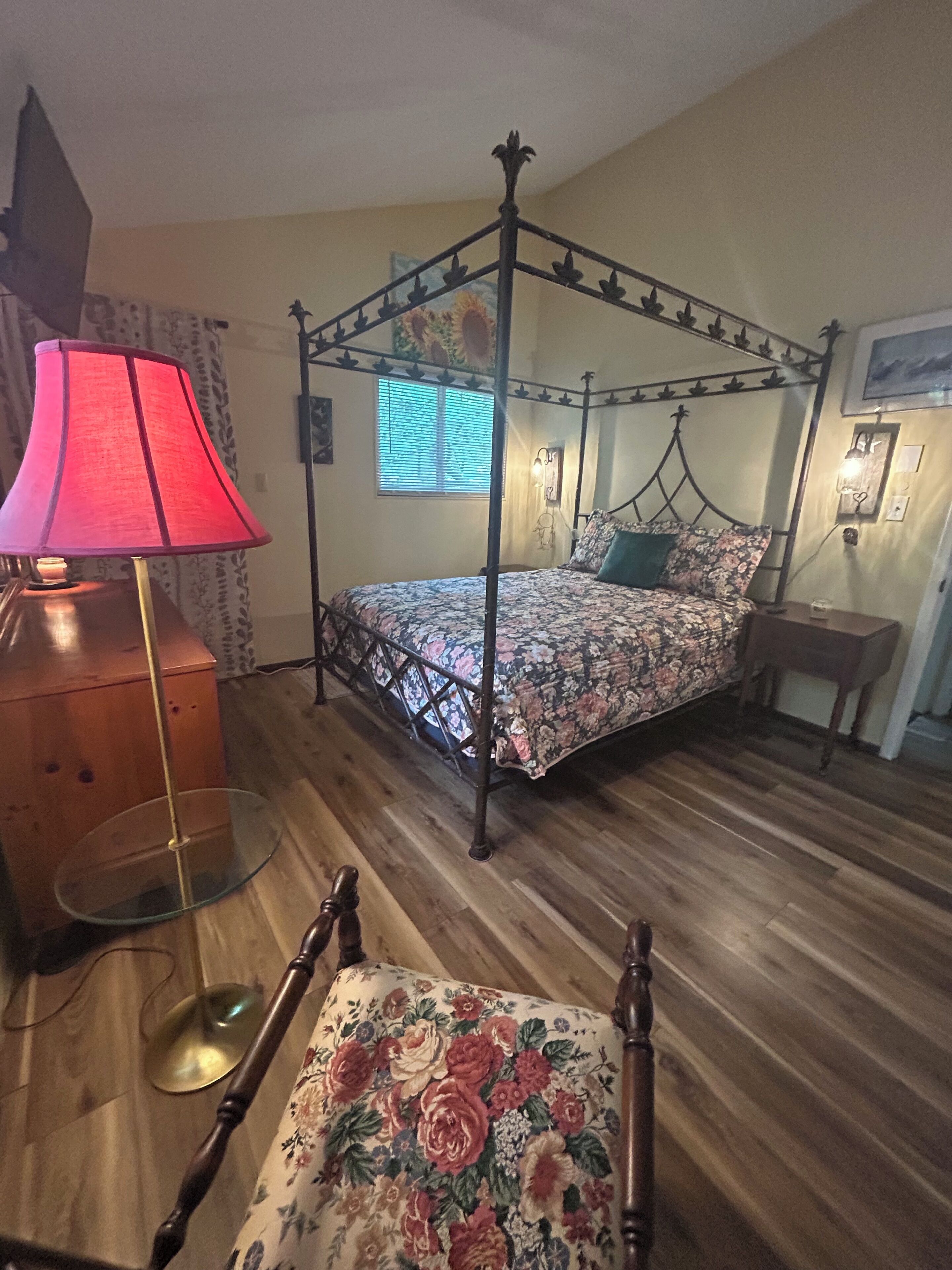 Un lit à baldaquin avec une literie à motifs floraux, une table de chevet en bois, une lampe rouge et une chaise recouverte d’un tissu à motifs floraux.