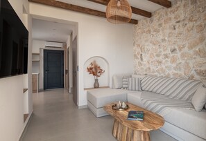 A modern living room with a stone wall, a sofa with patterned cushions, a wooden coffee table with books, and a hanging light fixture. - Strovilla Gaia 3 Luxury Private Beachfront Villas (Psakoudia)