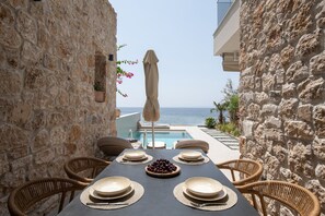 Une table à manger dressée pour quatre personnes, avec vue sur la mer, des murs en pierre et une piscine.