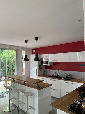A modern kitchen with white cabinets, a wooden countertop, and a red accent wall. There are two pendant lights, a stainless steel oven, and a sink with a dish rack. - Abatilles-Arcachonnaise, 10-person capacity - Near the beach and shops (Arcachon)