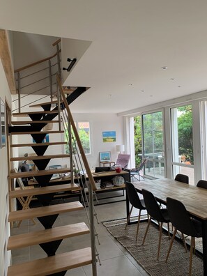 A modern interior with a wooden staircase, a dining area with a long table and chairs, and large windows offering a view of the outdoors. - Abatilles-Arcachonnaise, 10-person capacity - Near the beach and shops (Arcachon)