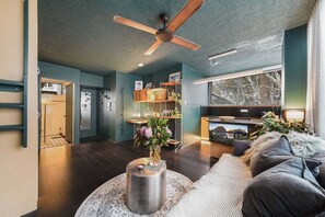 Ein modernes Wohnzimmer mit Deckenventilator, rundem Couchtisch mit Vase, Sofa und Küchenzeile mit Fernseher.