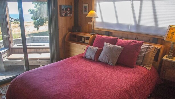 A bedroom with a large bed, a nightstand, a lamp, and a view of the outdoors. - Moon Shadow (TOT ID #2164): 2  BR, 2  BA House in The Sea Ranch, Sleeps 4 (The Sea Ranch)