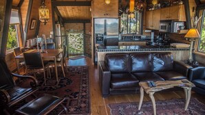 A cozy interior with a leather sofa, dining table, and chairs, a kitchen area with appliances, and a large window offering a view. - Moon Shadow (TOT ID #2164): 2  BR, 2  BA House in The Sea Ranch, Sleeps 4 (The Sea Ranch)