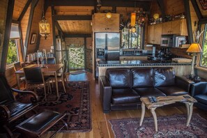 A cozy interior with a leather sofa, dining table, and chairs, a kitchen area with appliances, and a large window offering a view. - Moon Shadow (TOT ID #2164): 2  BR, 2  BA House in The Sea Ranch, Sleeps 4 (The Sea Ranch)