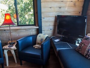 Een kamer met een televisie, een blauwe bank, een klein tafeltje met een lamp en een raam met uitzicht op bomen.