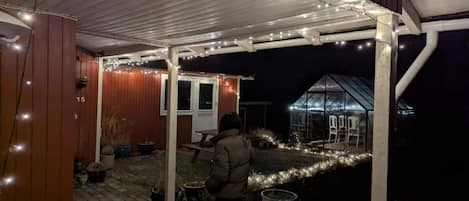 A covered outdoor area with a wooden structure, potted plants, and a greenhouse.