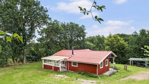 A red single-story house with a covered porch, surrounded by a grassy yard and dense trees. - 5 Star Holiday Home in Jaegerspris (Jægerspris)