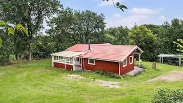 A red single-story house with a covered porch, surrounded by a grassy yard and dense trees.