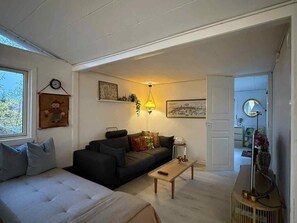 Sala de estar com sofá, mesa de centro e uma cama. Uma janela oferece vista para árvores.