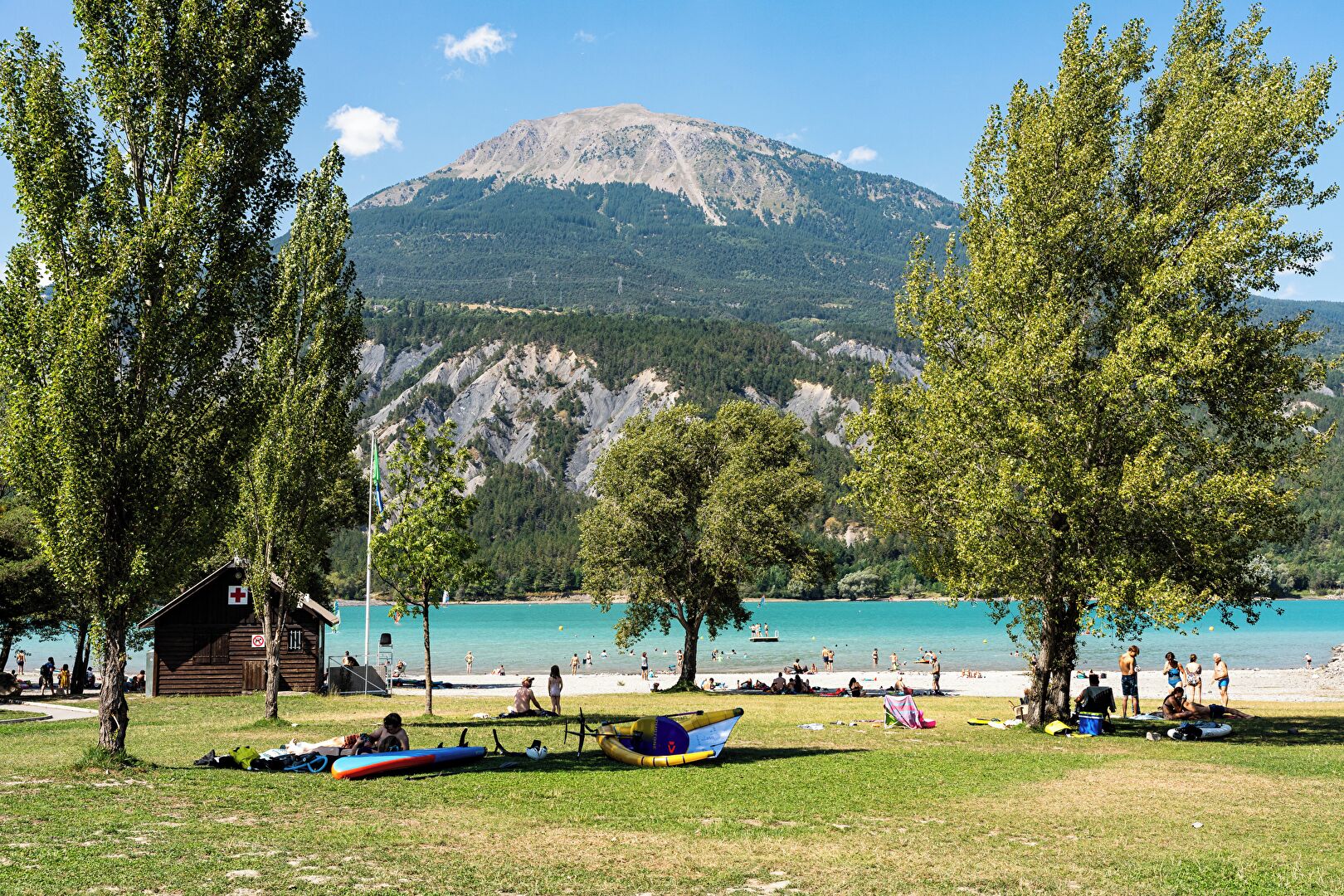 A lakeside view with a mountain in the background, a small wooden cabin, and several canoes on the grass.