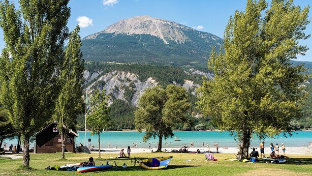 A lakeside view with a mountain in the background, a small wooden cabin, and several canoes on the grass. - Appartement 4 Personnes 31m² - Les Orres 1650 (Les Orres)