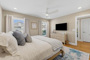 A bedroom with a bed, curtains, a ceiling fan, a television, and a view of the outside. - 🌊Sandman by the Sea - Pet Friendly 4 Bedroom Home with Spacious Decks🏖️ (Kure Beach)