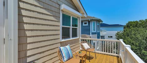 A wooden deck with two wicker chairs and a table, a white railing, and a window with shutters.