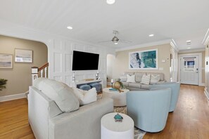 Ein großzügiger Wohnraum mit einem großen Flachbildfernseher, einem grauen Sofa, einem blauen Sessel und einem weißen Couchtisch.
