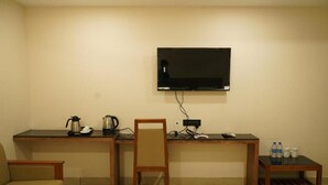 Ein Zimmer mit an der Wand montiertem Fernseher, einem Schreibtisch mit Stuhl, einer Kaffeemaschine und einem kleinen Tisch mit Wasserflaschen.
