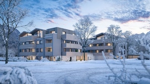 Un immeuble moderne à plusieurs étages, doté de balcons, entouré d’arbres et de montagnes couverts de neige.