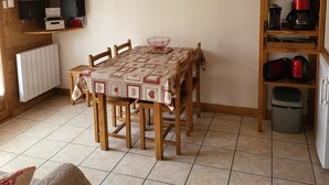 Ein gemütlicher Essbereich mit Tisch für vier Personen, einem Fernseher auf einem Holzregal, einem gerahmten Bild an der Wand und einer Küchenzeile mit Wasserkocher und Kaffeemaschine.