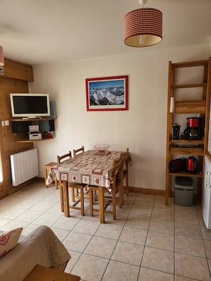 A cozy dining area with a table set for four, a television on a wooden shelf, a framed picture on the wall, and a kitchen area with a kettle and coffee maker. - 3-star apartment at the base of the slopes, large terrace, 2 bedrooms (Les Deux Alpes)