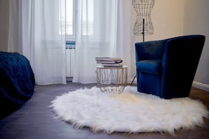 Una habitación con una silla azul, un tapete blanco y mullido, y una pila de libros sobre una mesita redonda.