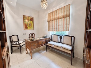 A room with a wooden desk, two chairs, a sofa, a window with blinds, a chandelier, and a painting on the wall.