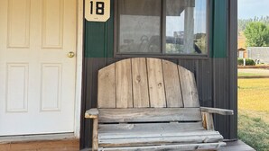 Un banc en bois se trouve devant un bâtiment. On peut y voir une porte portant le numéro « 18 » et une fenêtre avec des stores.