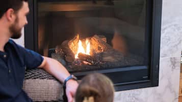 Une cheminée avec des flammes apparentes, une personne assise sur un canapé et une autre personne assise sur le sol.
