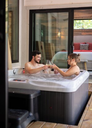 Ein Whirlpool mit zwei Personen, die mit Gläsern anstoßen, eine Holzterrasse und eine Glastür zu einem Außenbereich.