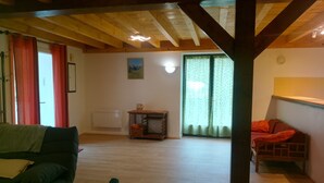 Um quarto espaçoso com teto de madeira, um sofá verde, uma mesinha com um jarro e vista pela janela com cortinas verdes.