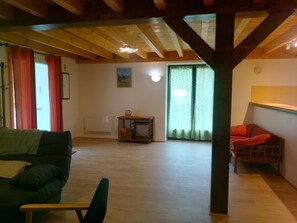 Ein geräumiges Zimmer mit Holzbalkendecke, einem grünen Sofa, einem kleinen Tisch mit einer Karaffe und Blick durch ein Fenster mit grünen Vorhängen.
