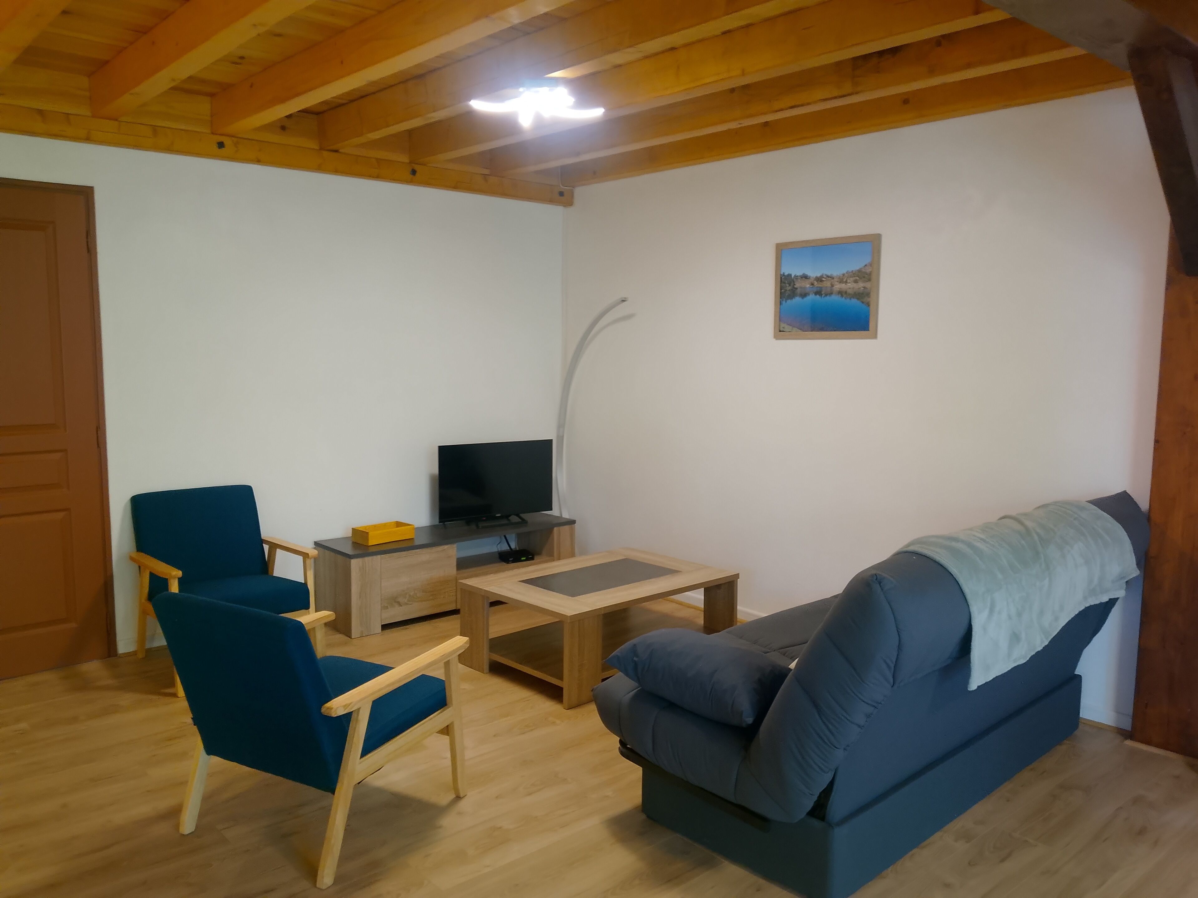A room with a wooden ceiling, a blue sofa, a wooden chair, a small table with a television, and a painting on the wall.