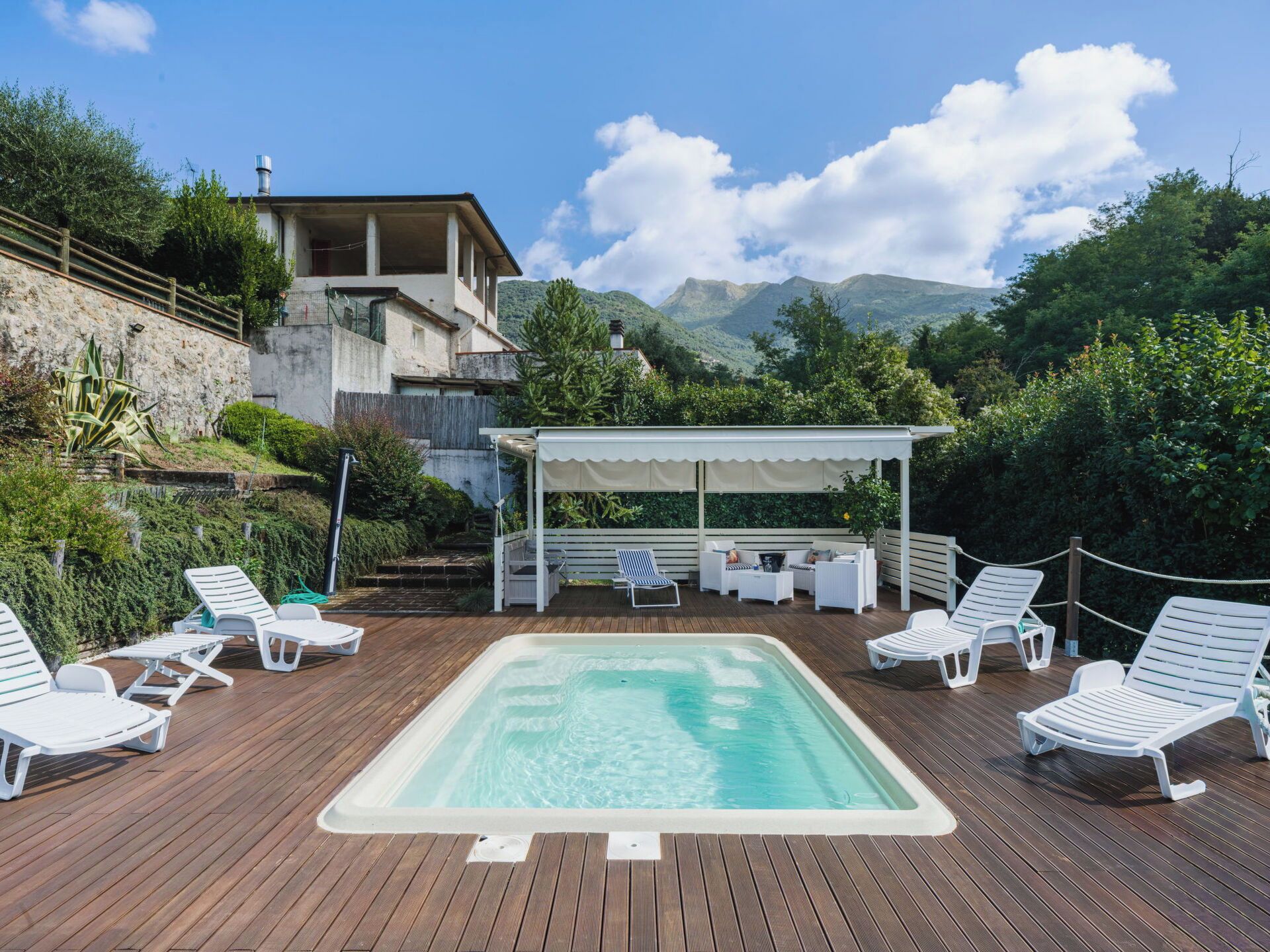 Une terrasse en bois avec un jacuzzi, entourée de chaises longues blanches, un espace salon couvert et une vue imprenable sur les montagnes.