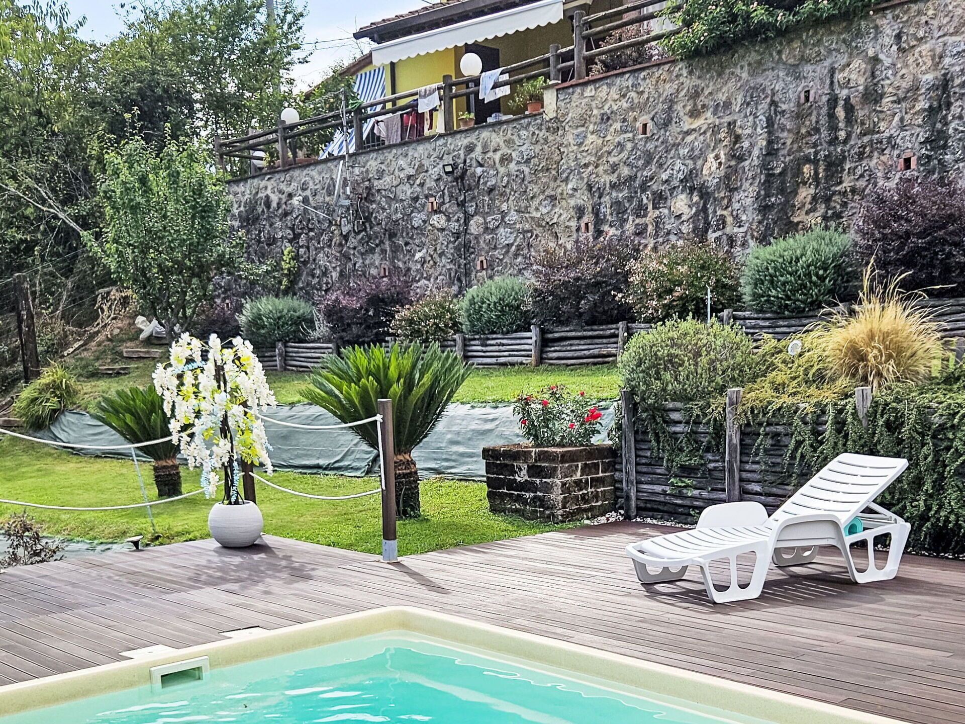 A pool area with a wooden deck, a lounge chair, and a stone wall with a balcony.