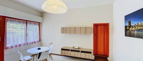 Un salon moderne avec un coin repas, une porte rouge et un tableau représentant un paysage urbain.