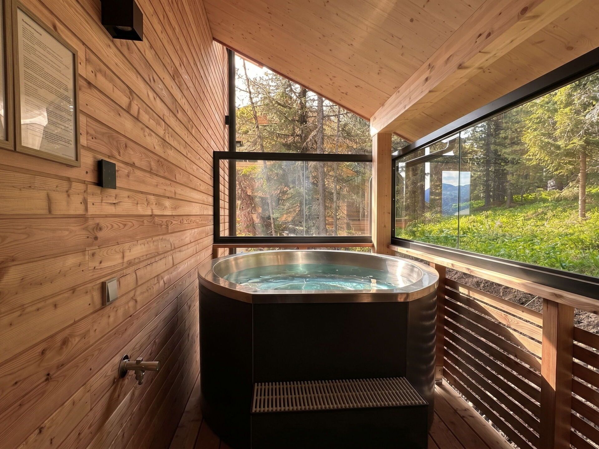 Interior de una cabaña de madera con jacuzzi, paredes de madera y un amplio ventanal con vistas al bosque.