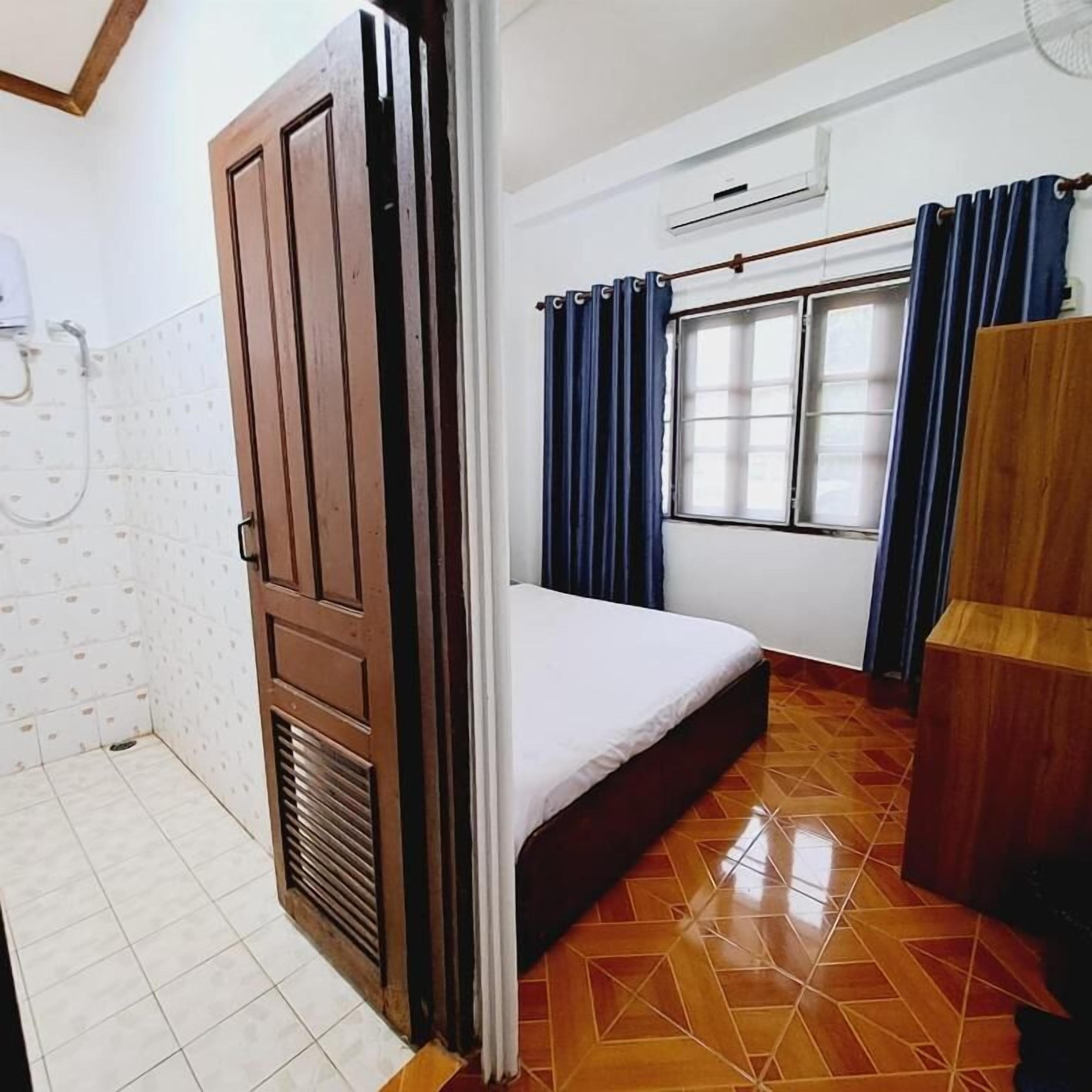 Una habitación con cama, ventana con cortinas, armario de madera y suelo de baldosas.