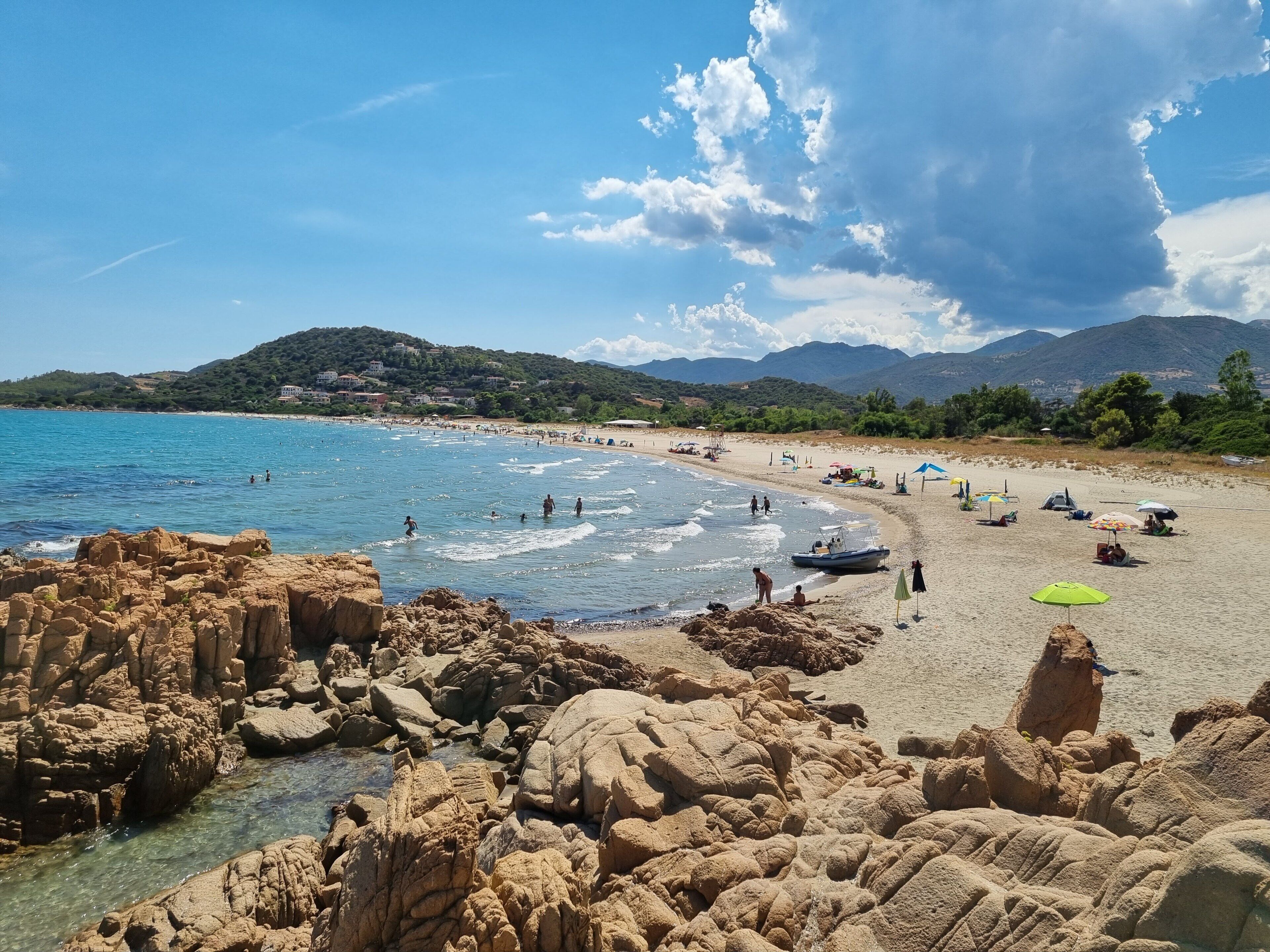 A beach with rocky formations in the foreground, a sandy beach area with umbrellas and people, and mountains in the background.